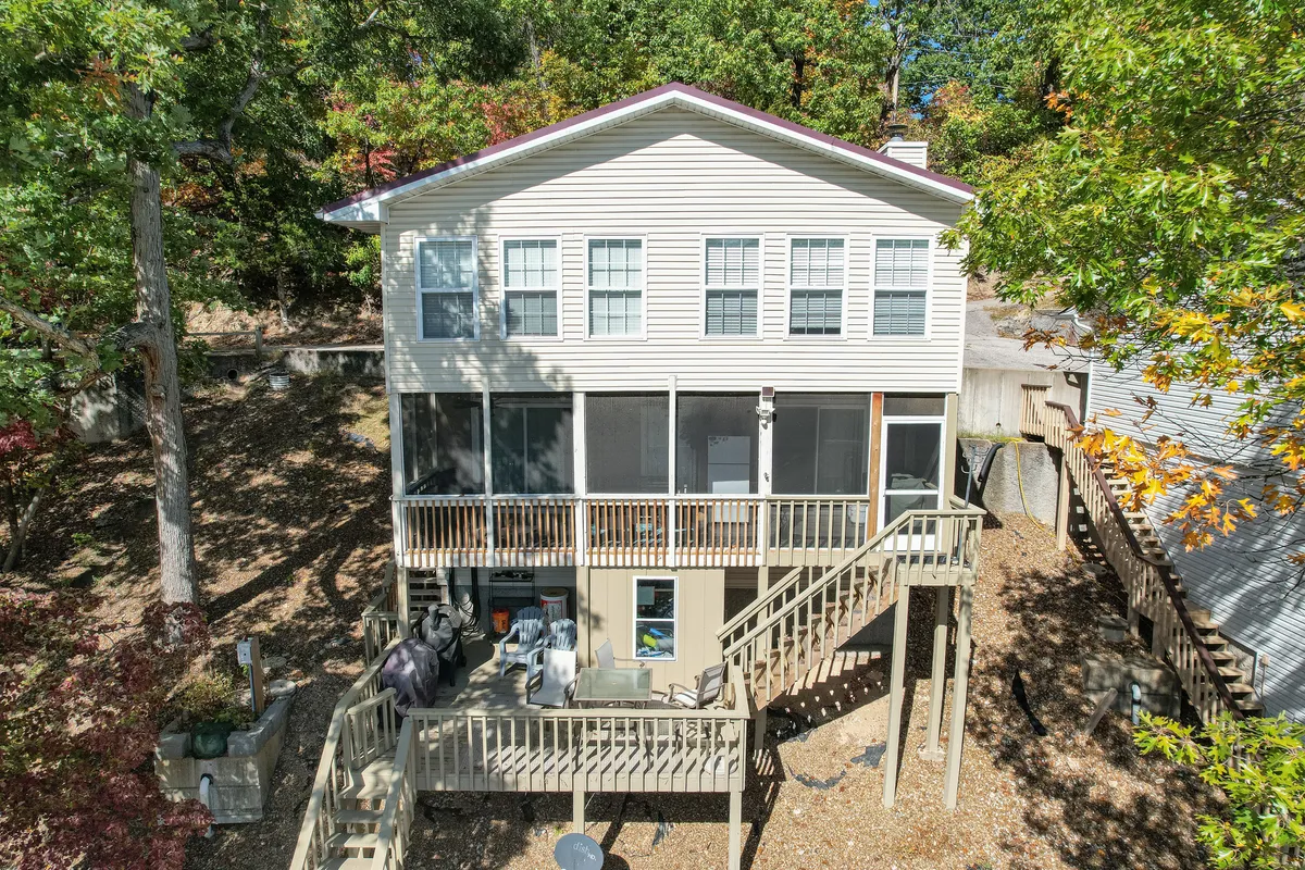 This image features a screened porch with a wooden deck and a staircase leading down, surrounded by lush trees. The setting exudes tranquility and invites viewers to imagine relaxing or gathering in a shaded, breezy space while still feeling closely connected to nature and the lake just beyond.