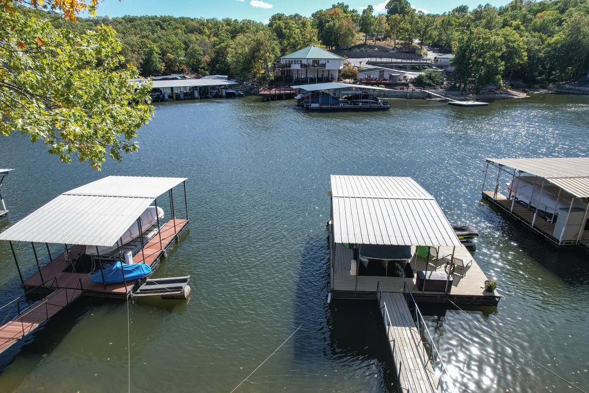 A serene dock with boat slips extends over the calm lake, surrounded by lush green trees and with houses visible on a gentle hill in the distance. The tranquil water reflects the evening sky, evoking a peaceful lakeside atmosphere ideal for sunset gatherings or quiet reflection.
