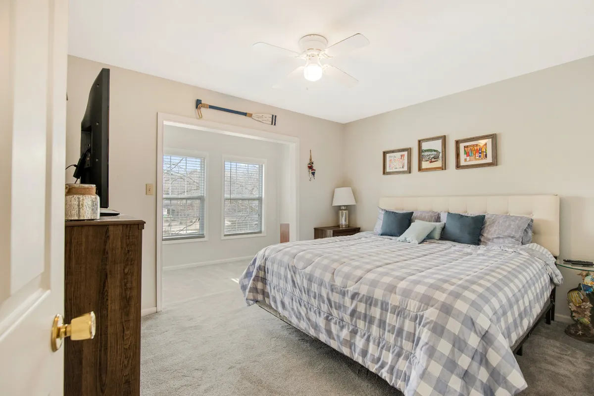 A welcoming bedroom featuring a bed with checkered bedding, large windows allowing natural light to pour in, and tasteful decorative wall frames. A nightstand with a lamp completes the cozy, restful setting, perfect for quiet mornings or evening relaxation.