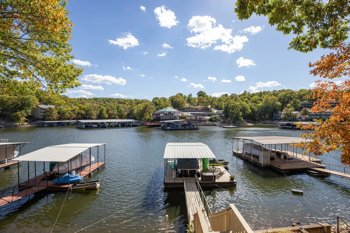A sweeping lakeside view highlights the sparkling water framed by boathouses, tall trees, and drifting clouds. The serene setting invites guests to imagine boating, fishing, wildlife spotting, or simply relaxing by the shore throughout the changing seasons. The peaceful mood captures the essence of lakeside leisure and retreat.