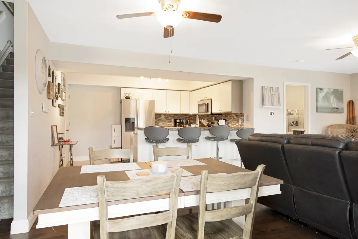 A spacious interior scene including a dining table, a kitchen island with stools, and a cozy living room seating area in the background. The space is filled with natural light, and the design encourages gatherings for meals, conversation, and casual entertaining.