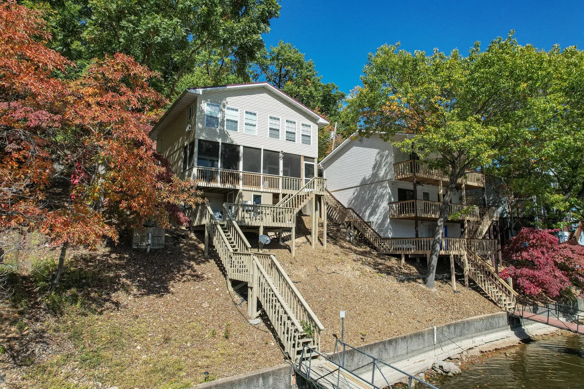 This exterior shot features two charming lake houses surrounded by lush trees, with a staircase leading down towards the tranquil waters of the lake. The spacious porches are visible, evoking scenes of outdoor relaxation, gatherings, and evening barbecues. The natural setting highlights the property’s direct connection to the lake and the beauty of the surrounding landscape in any season.