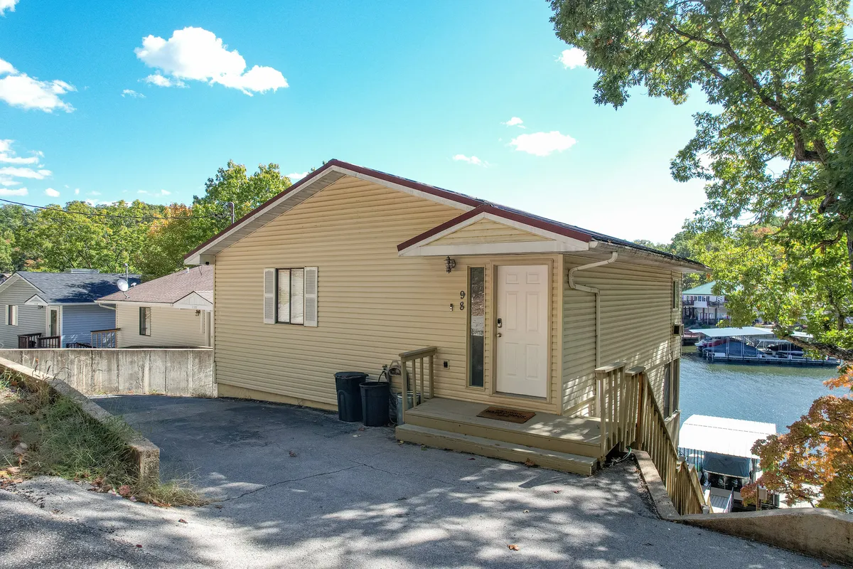 An exterior shot featuring the front door, deck, landscape, and visible boat docks. The welcoming scene shows spaces for outdoor gatherings, barbecues, and immediate lake access.