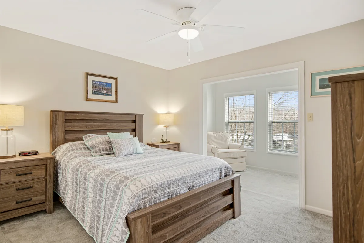 A main bedroom with a plush bed adorned in patterned bedding, flanked by nightstands with glowing lamps. The space is bathed in natural light streaming through large windows, creating a tranquil, cozy atmosphere ideal for relaxation or reading.