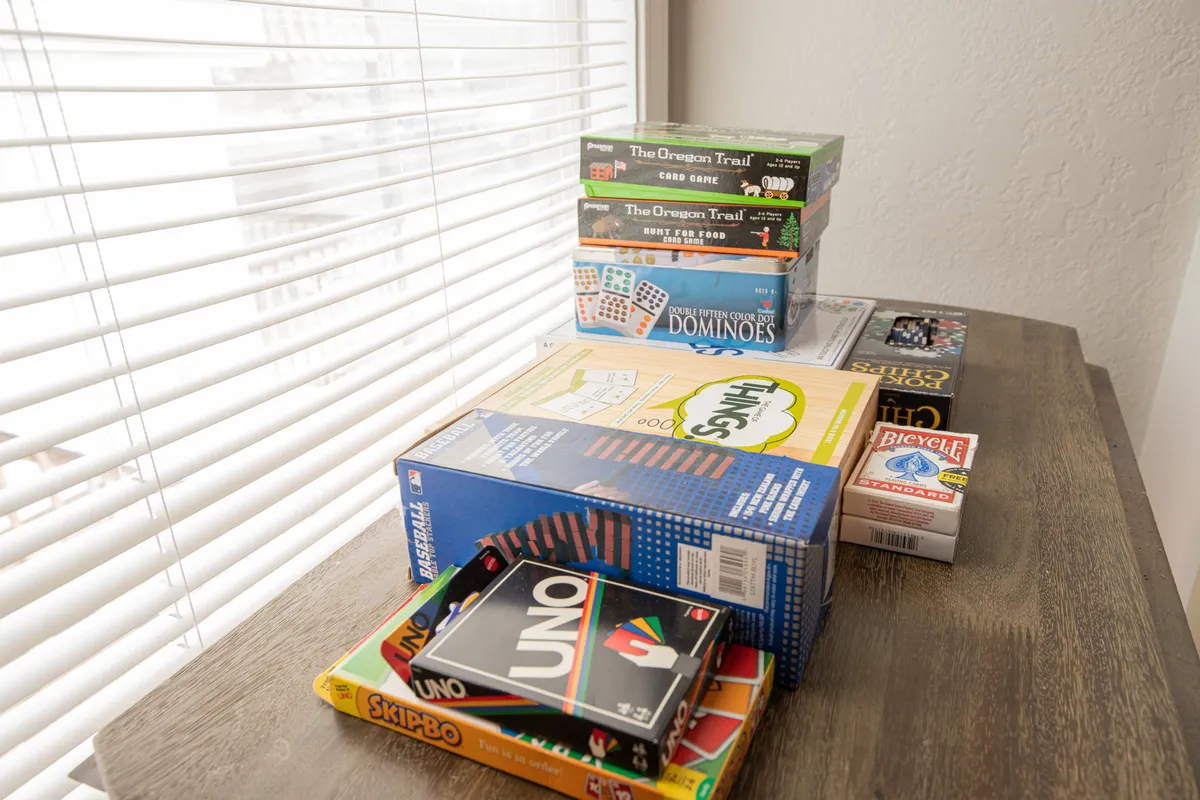 An interior shot focused on a wooden table stacked with a variety of board games, set beside a window with natural light. The playful setup suggests casual leisure and family bonding, complementing the cozy indoor activities described for colder or quieter months.