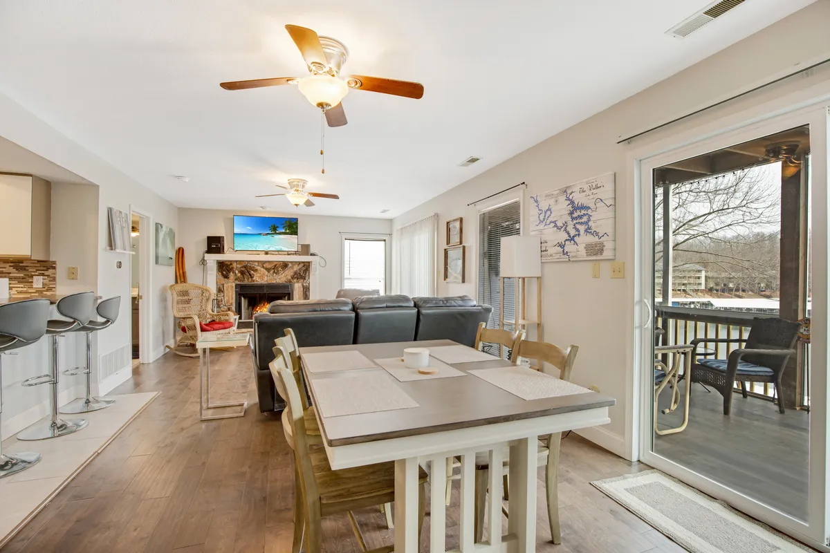 A welcoming open-concept living and dining area featuring a central dining table, a plush sofa facing a fireplace and television, and a large sliding door that frames the patio and brings in natural light. The setting highlights both communal and relaxation spaces, with soft furnishings and a bright, airy atmosphere.