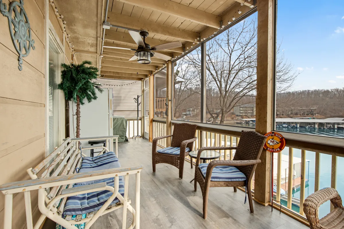 A peaceful screened-in porch with wicker chairs, wooden railing, ceiling fan, and lush greenery framing a direct view of the lake. The scene suggests a shaded spot for relaxing, enjoying a snack, or unwinding together while still feeling connected to the outdoors.